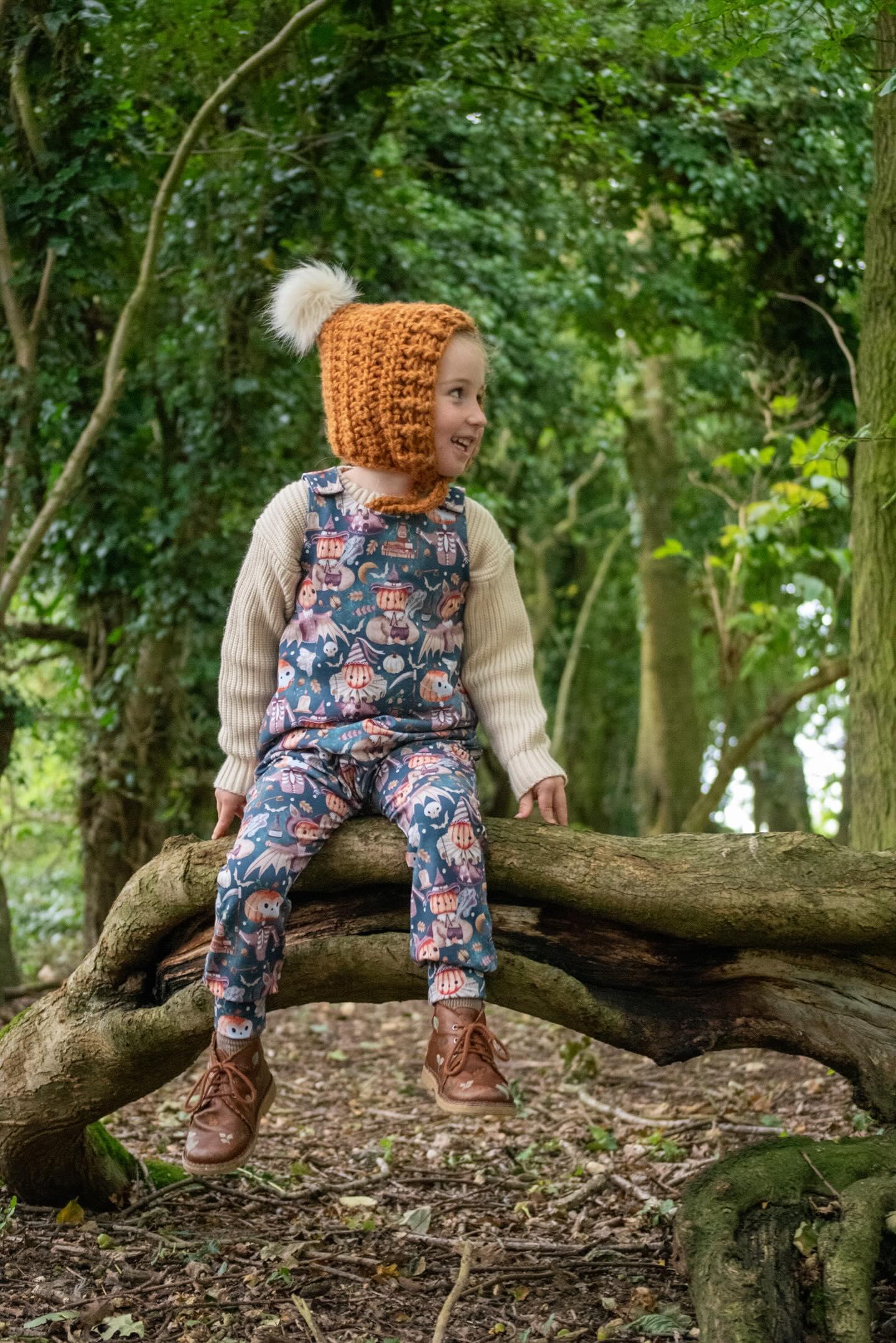 Boy in a tea and crochet hat, cream jumper and Halloween pumpkin romper sat on a tree stump